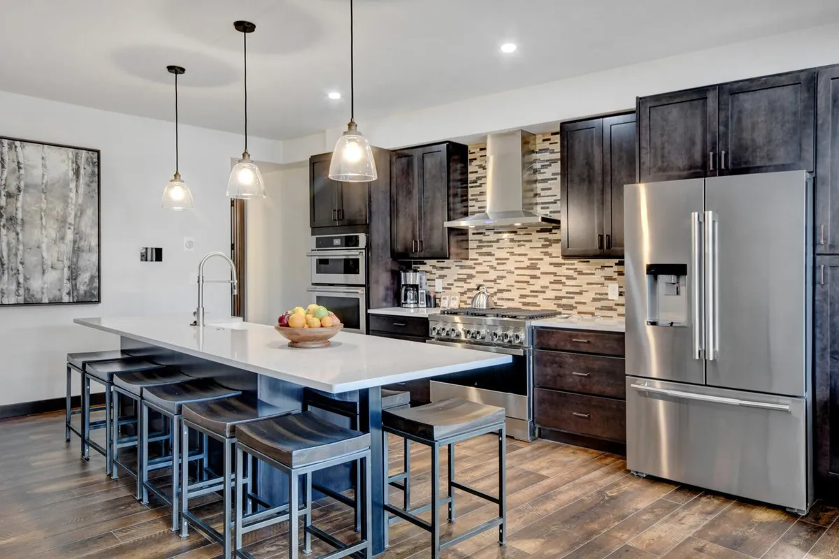 This image features a modern kitchen with a central island and bar seating, stainless steel appliances, a textured backsplash, and pendant lighting. The scene conveys a sense of togetherness and culinary creativity with ample room for guests to gather and mingle while meals are being prepared.