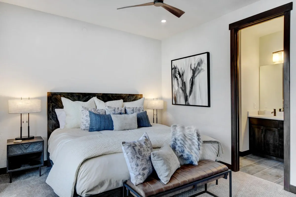 A tranquil king suite with a king-sized bed adorned with decorative pillows, soft bedding, and nightstands with lamps. Artwork adds a stylish touch, and an entrance to a private bathroom is visible. The atmosphere is restful and cozy, inviting relaxation and peaceful sleep.