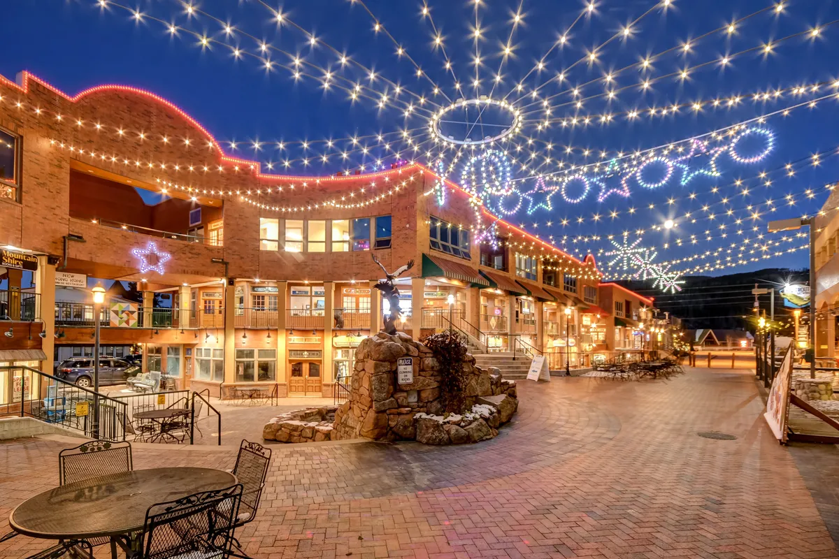 A lively outdoor scene featuring string lights overhead, decorative star accents, a stone fountain, and comfortable outdoor seating. People can be imagined socializing, dining, or celebrating beneath the festive lights, with boutique shops and eateries lining the area.