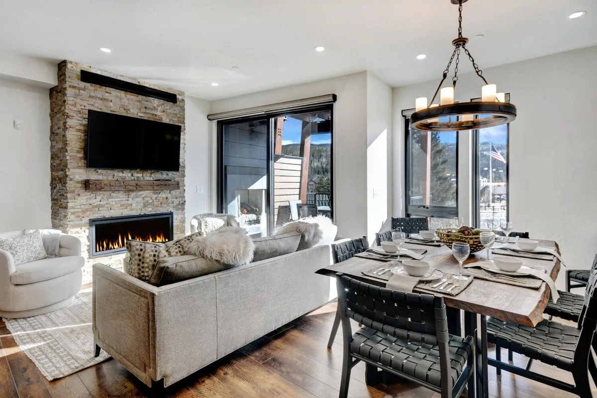 A welcoming open-plan living and dining space featuring a glowing fireplace, a large wooden dining table, plush sofa, and expansive windows framing natural light. Cozy throws are draped over the seating, and the ambiance is warm and inviting—ideal for relaxing after outdoor adventures or gathering with family and friends.