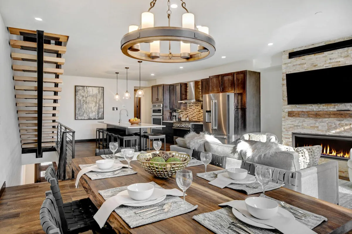 This image showcases a stylish open-concept living and dining area with a grand dining table set for guests, a large kitchen island, a glowing fireplace, and a beautiful chandelier overhead. The space is warmly lit, inviting, and clearly designed for gatherings and entertaining.