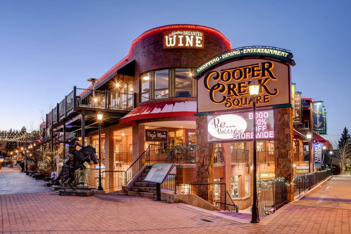 An inviting exterior scene of Cooper Creek Square, with its decorative lighting, colorful signage, outdoor seating, and a horse sculpture, bustling with activity. It evokes the festive, welcoming atmosphere of downtown Winter Park, with shopping, dining, and entertainment close at hand.