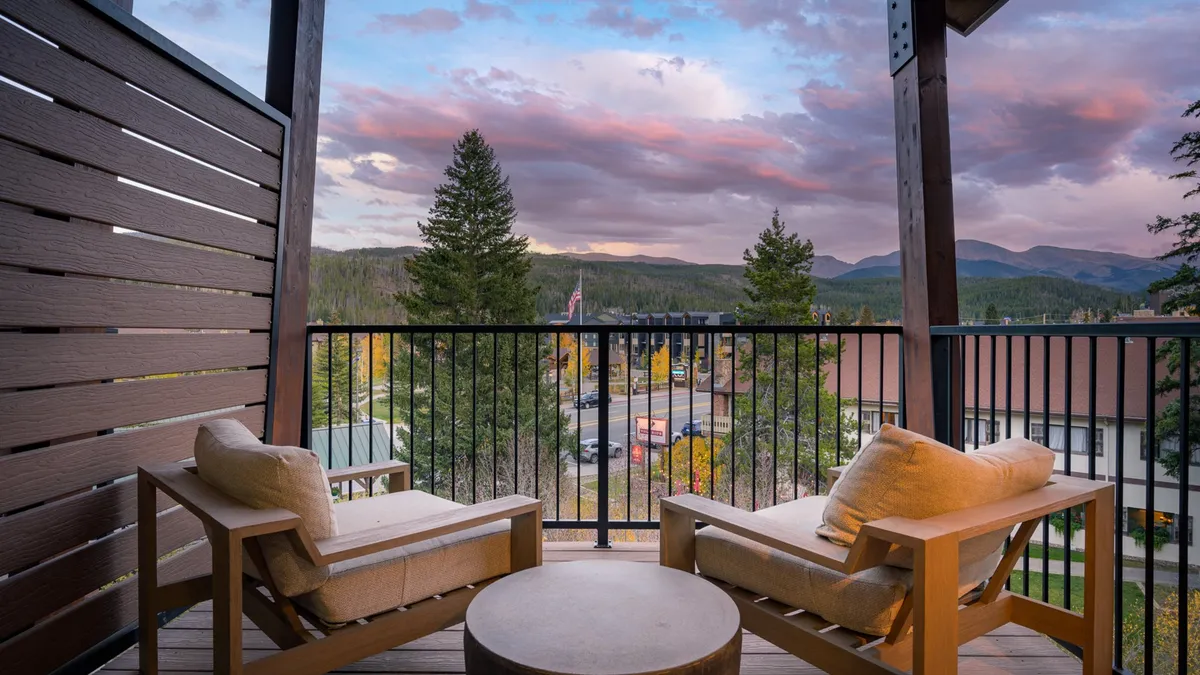 A relaxing patio scene with rocking chairs and a small table, set against a panoramic backdrop of mountains and dramatic skies. The inviting setup evokes evenings spent watching sunsets, stargazing, or enjoying patio dining in the summer.