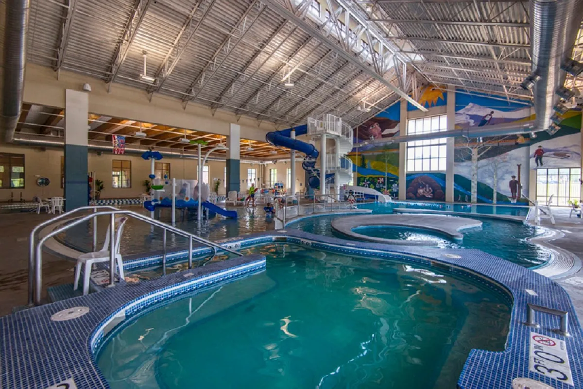 A vibrant indoor recreation center with children gleefully playing in a swimming pool and zipping down colorful water slides. The space is lively, adorned with a playful mural, and brimming with family-friendly activity.