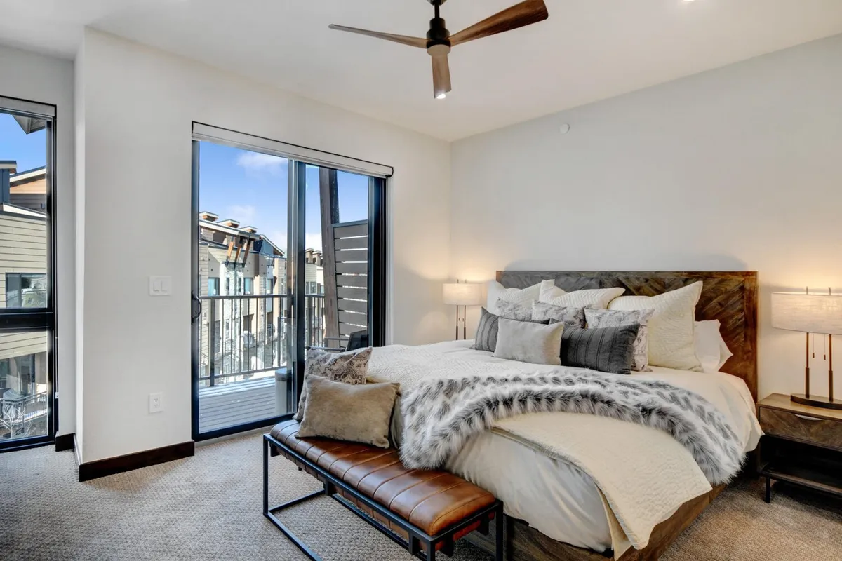 A serene bedroom featuring a large bed with decorative pillows, nightstand lamps, a textured throw blanket, and a bench at the foot of the bed. Sliding doors open to a balcony with views, letting in abundant natural light.