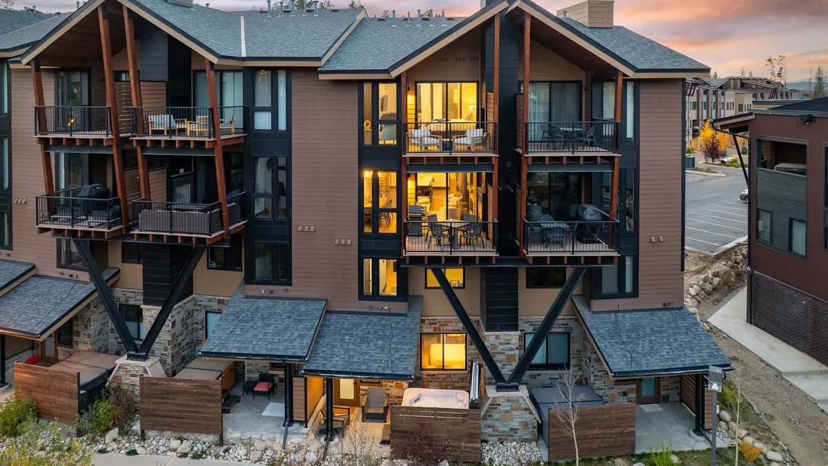 The image features the exterior of a luxurious single-family villa set along a river in Winter Park, Colorado. Spacious balconies with comfortable seating overlook scenic vistas, with large windows reflecting the mountain landscape. Stone accents give the home a refined, rustic elegance. The scene suggests guests relaxing on balconies, enjoying sunset views, and gathering for outdoor meals or morning coffee.