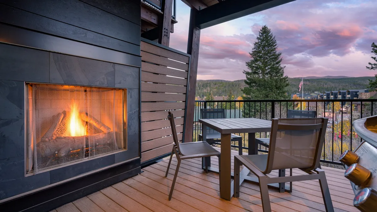 An outdoor seating area set around a stone fireplace with comfortable chairs and a table, all overlooking a sweeping mountain view beneath a dramatic sky. The scene suggests moments spent dining al fresco, sharing warm drinks, or stargazing by the outdoor fire.